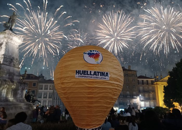 Um balão de papel laranja com o logotipo 'Huellatina' flutua sobre uma praça movimentada, com fogos de artifício iluminando o céu noturno.