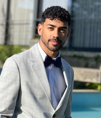 Pablo Jovem homem com cabelo encaracolado e barba, sorrindo e usando um terno cinza xadrez e uma gravata borboleta azul marinho.